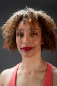 A multiracial Black woman with coffee-colored skin and short, curly hair. She smiles softly at the camera; her crimson lip color and coral tank top pops.