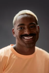 Jerron Herman headshot. He is a dark-skinned Black man with a big smile, dark moustache, and bright blonde hair; he wears a soft yellow T-shirt.