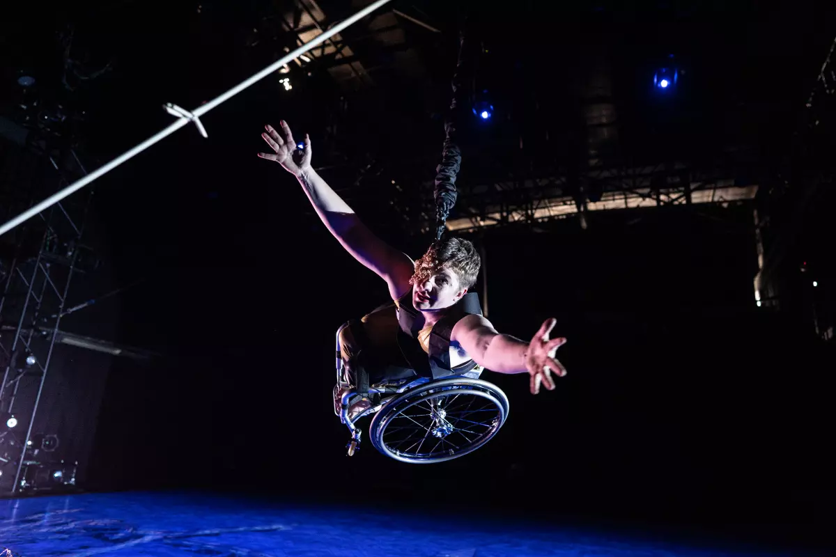 Laurel Lawson, a white woman with very short cropped hair, bounds toward the camera: wheelchair high off the ground, arms open and reaching. A mask of delicate gold wire and lustrous pearls covers half her face. The stage floor below her is drenched in blue light; the same light gives her pale skin a light violet glow. A strand of silver barbed wire appears, close up, in the upper corner.