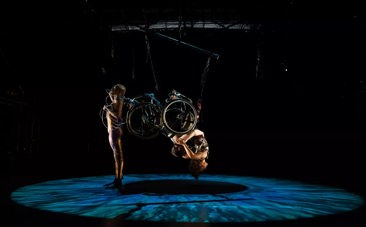 Three dancers appear onstage in a ring of dappled green and blue light. Jerron, a dark-skinned Black man with blonde hair, stands straight; he is wrapped in barbed wire, one arm resting on his chest, with his back to Alice and Laurel. Alice and Laurel hang upside down; their bodies and chairs entangled and becoming one. Laurel, a white woman with cropped hair, wraps her arms around Alice, a multiracial Black woman with coffee-colored skin and short curly hair. Alice arches and reaches her arms back, resting them on Laurel’s shoulders.