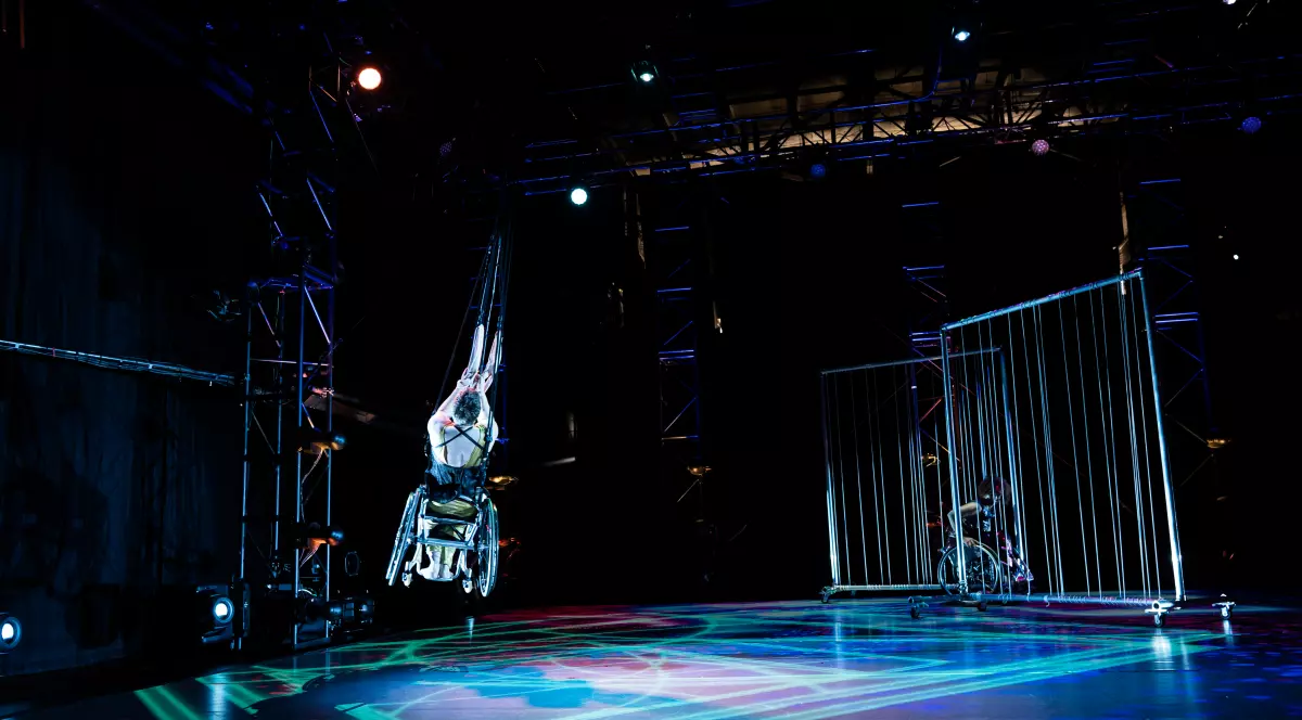 Laurel Lawson, a white dancer with short cropped hair, launches into the air; her back is to the camera as she pulls herself up, hands gripping loops and cables. Her gold costume, pale skin, and wheels shine in the light, in contrast to the dark stage. Another dancer appears in the distance, in shadow, wheeling behind tall wire gates. Cool green and blue patterns appear on the stage below.