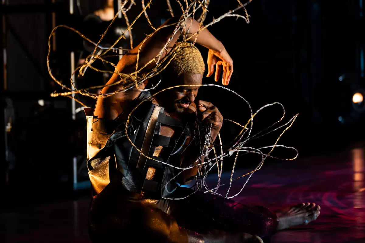 Jerron Herman, a dark-skinned Black man with blonde hair and a dark mustache, sits on the stage curled into a taut spiral; he is wrapped in a tangle of barbed wire. He wears a shimmery bodysuit and black leather top. His eyes are closed, a tense expression on his face; one arm arcs over his head, palm open, as the other is tucked at his shoulder in a fist