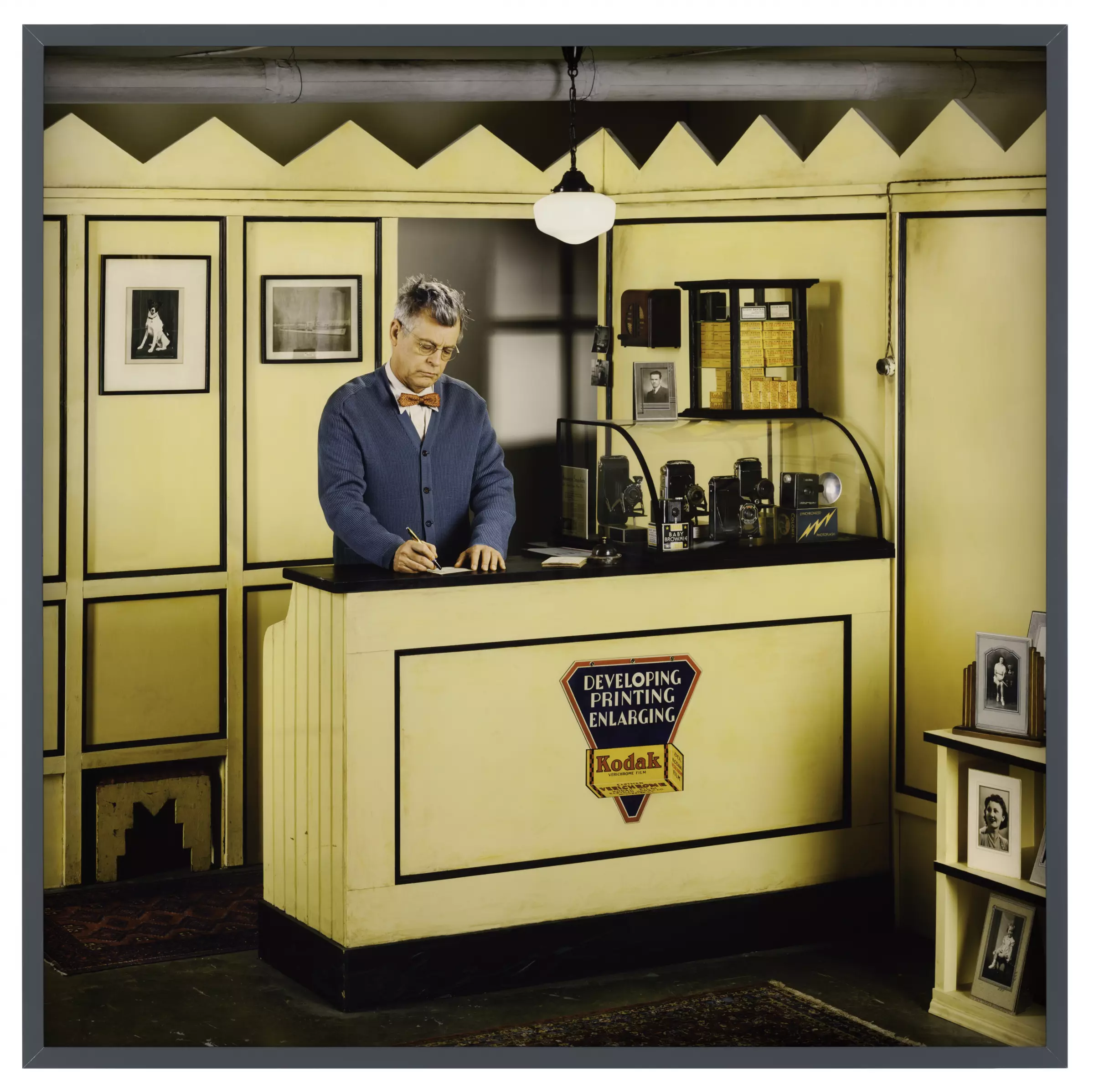 A man in a cardigan and bowtie stands behind the counter of a small booth. A Kodak advertisement reads 'developing printing enlarging'