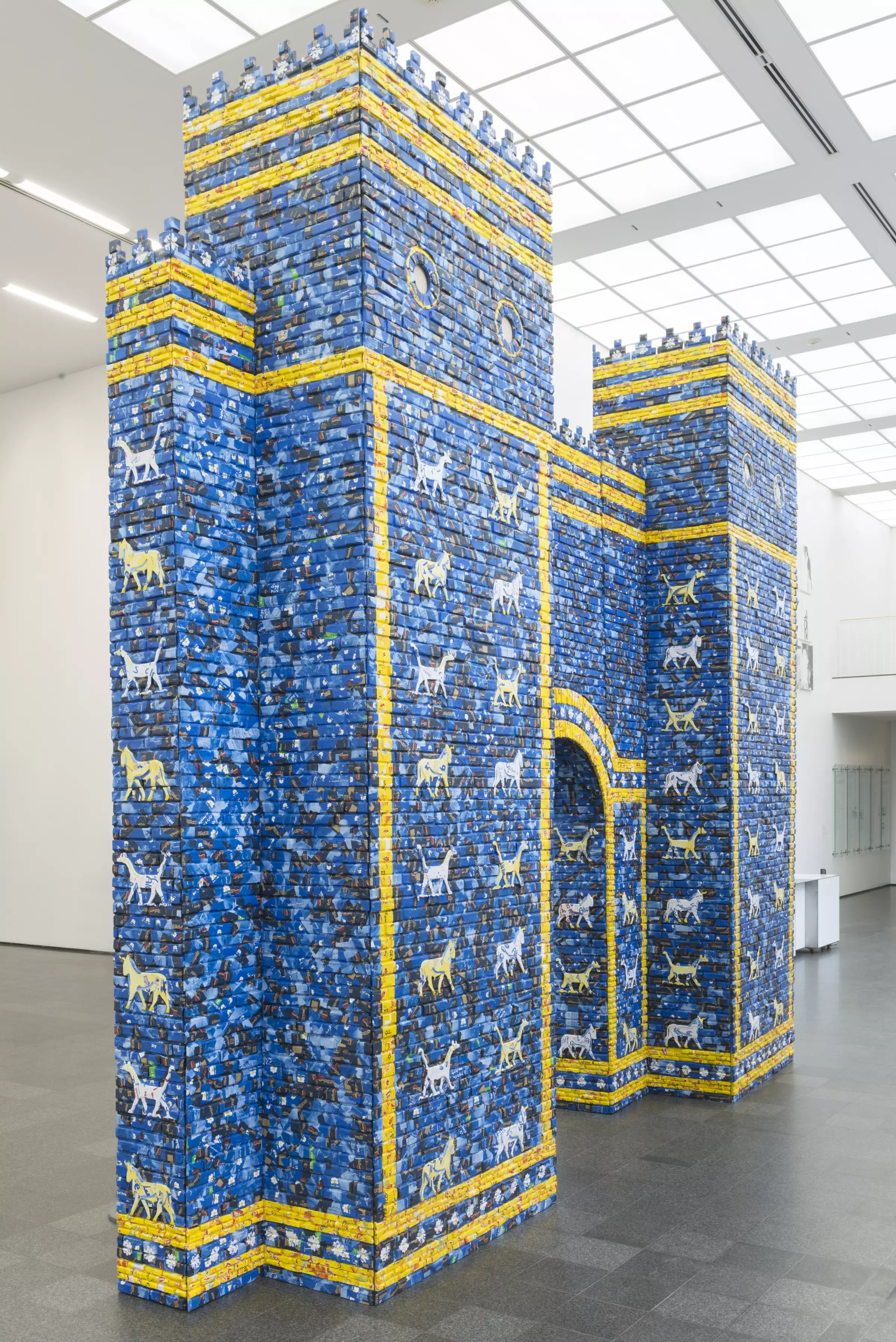 Installation view of a castle facade with crenelations, an arched doorway, and covered with blue tiles with yellow tile–stripes and columns of painted animals.