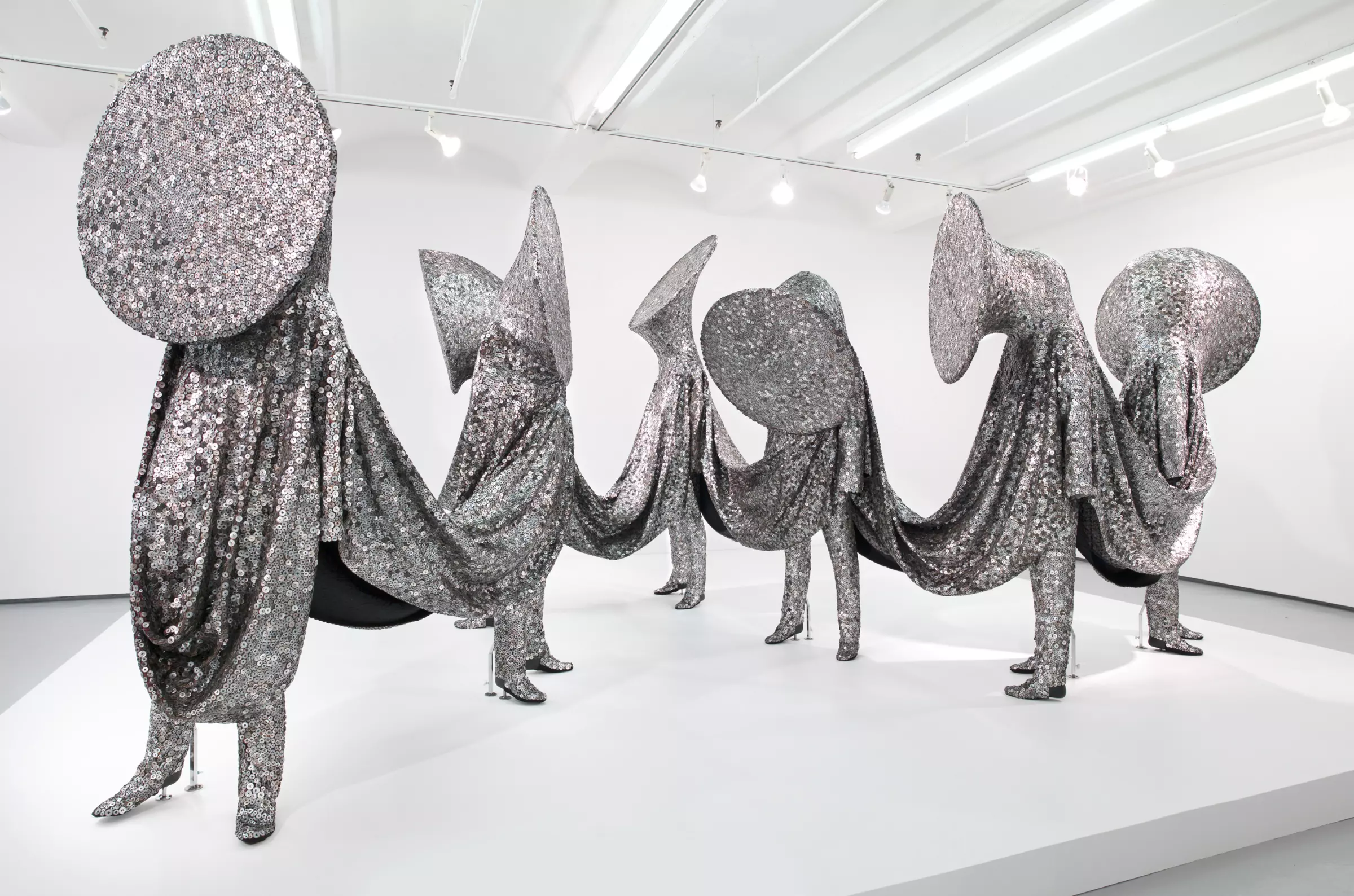 several figures with tuba-like heads and covered from head to toe in a metallic fabric stand on a white platform
