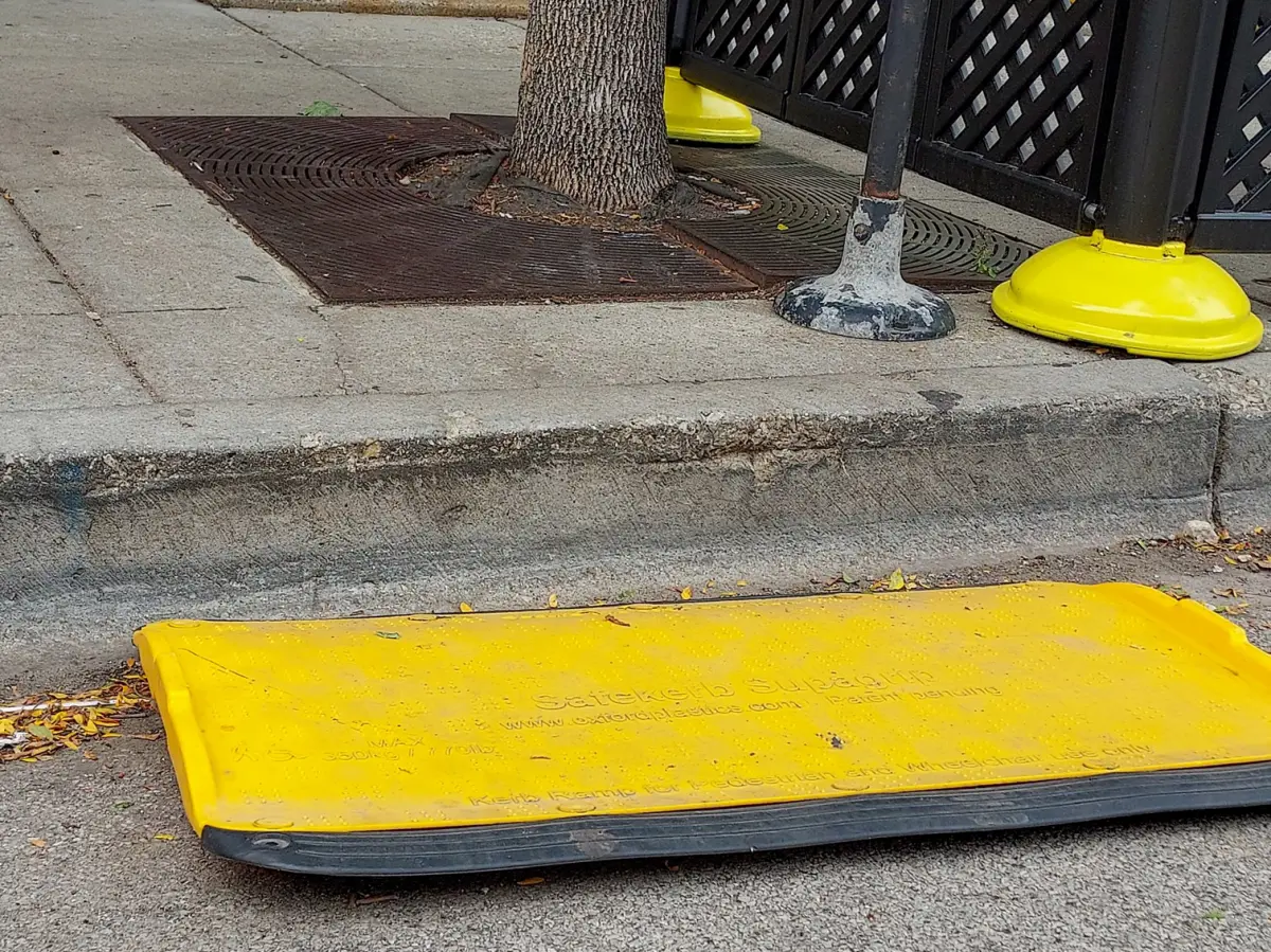A scuffed concrete city curb, a yellow plastic accessible ramp, and the distance between the two. Beyond the curb is a sidewalk, with a tree trunk, and the edge of a fence.