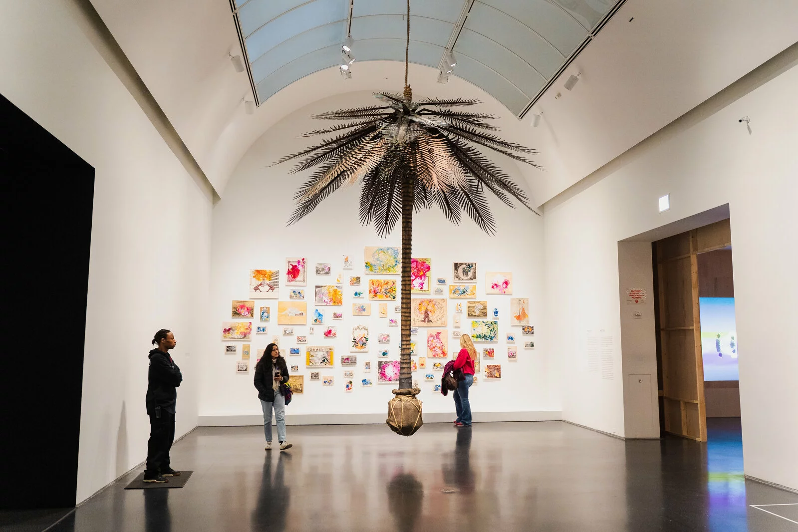 Three people stand around a palm tree hanging from the ceiling in a large room. Behind them are many colorful artworks hung on the wall in a gallery-style fashion.