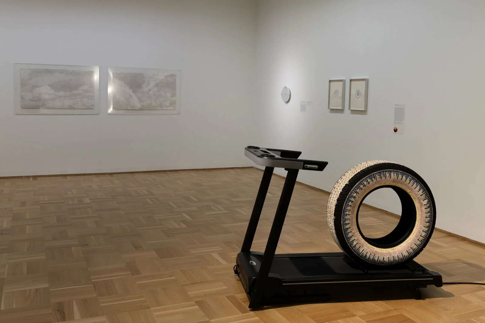 Gallery space with tire on a treadmill filling right foreground of image. Other artworks can be seen hung on the walls.