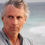 Close up portrait of a grey-haired man looking off to our left against a blurry ocean background.