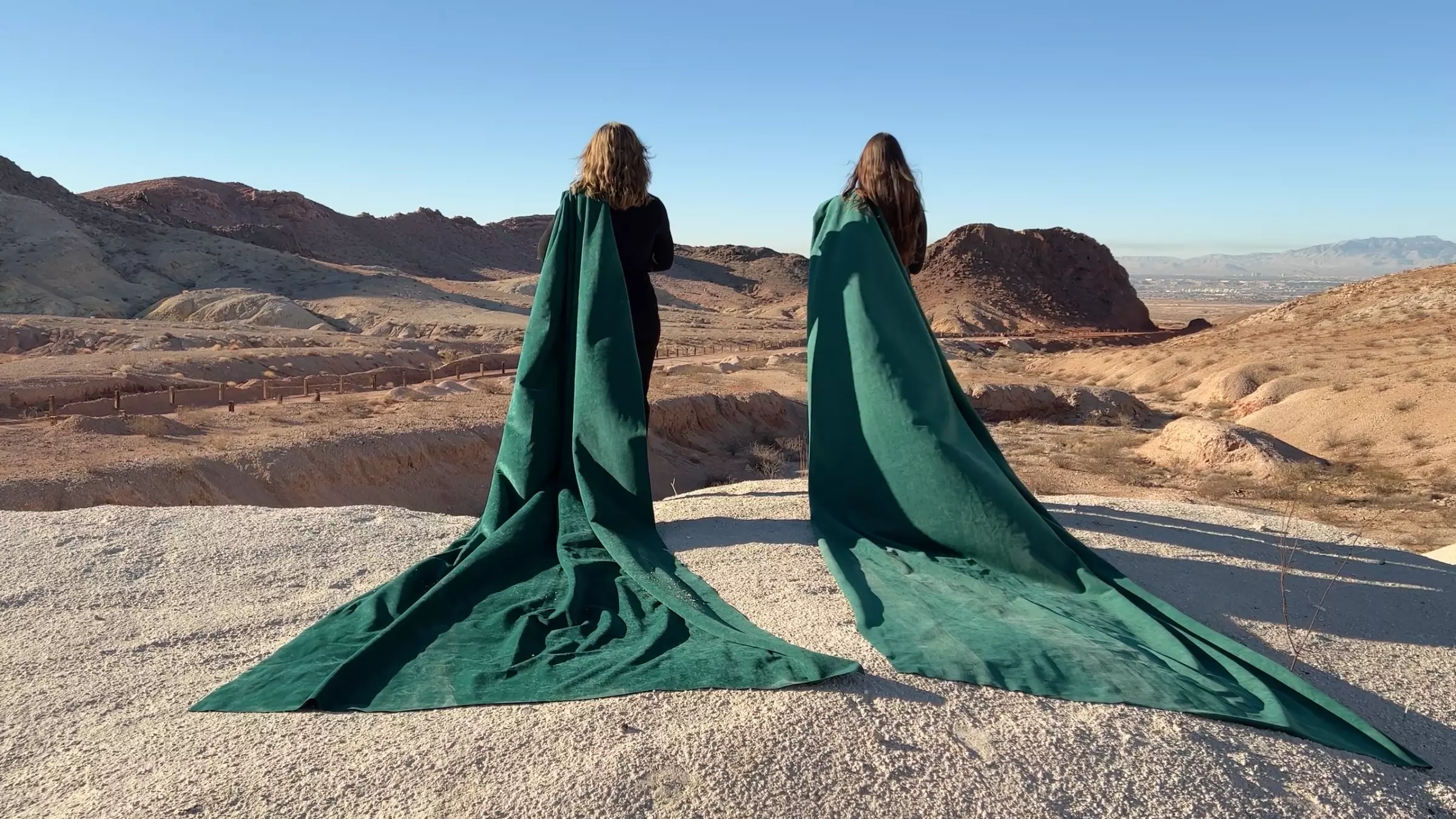 photograph of two people draped in long green capes facing a mountainous desert landscape