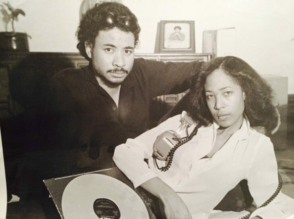 A Black man and woman are reclining with a record in front of them; the woman also has headphones.