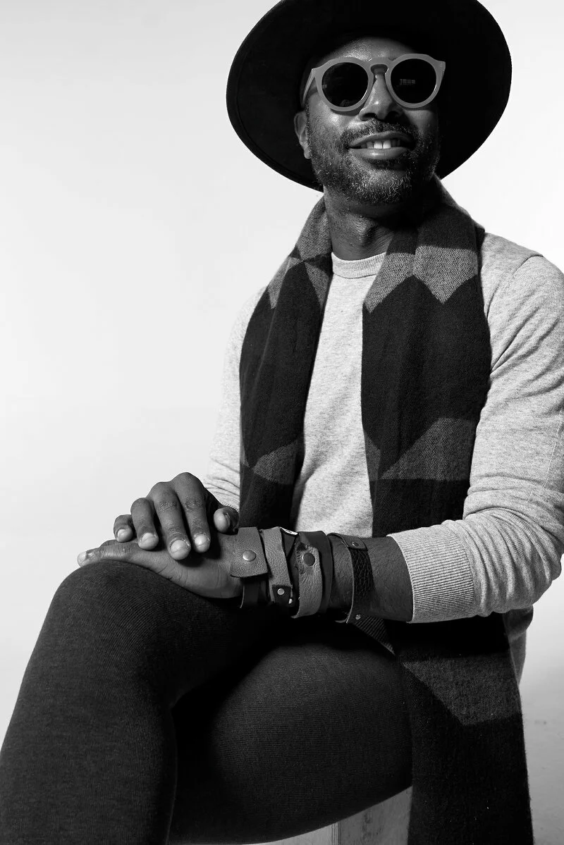 A Black man wearing a fedora-type hat, dark sunglasses, and a scarf sits with his hands folded and smiles, slightly off camera.