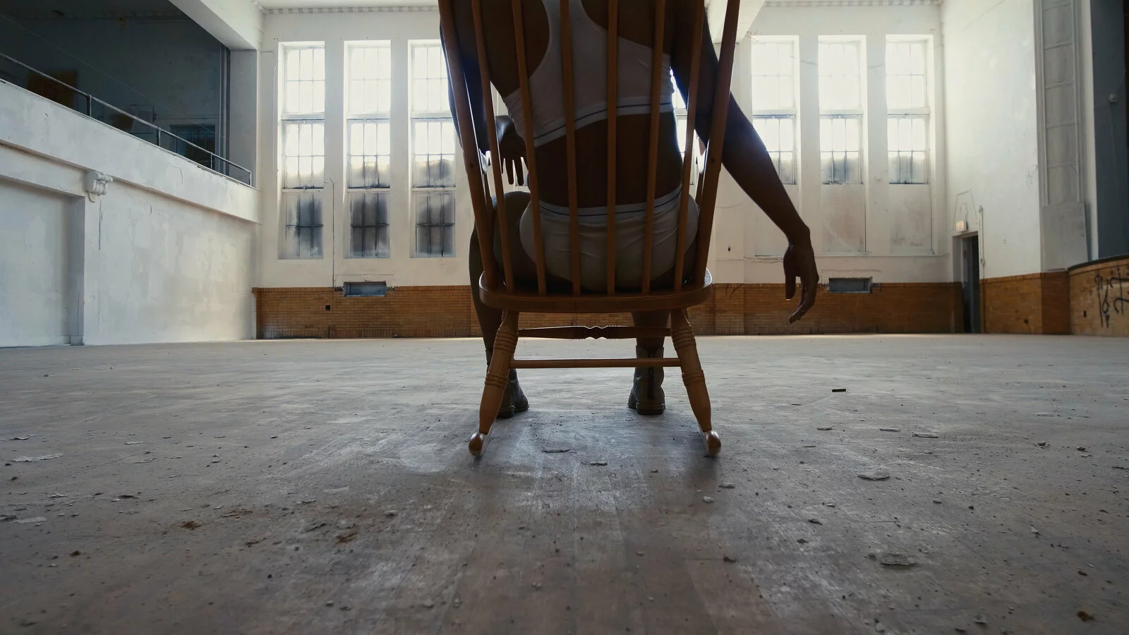 Wide-angled shot of a person, pictured from the shoulders down, sitting in a wooden chair in an otherwise empty room.