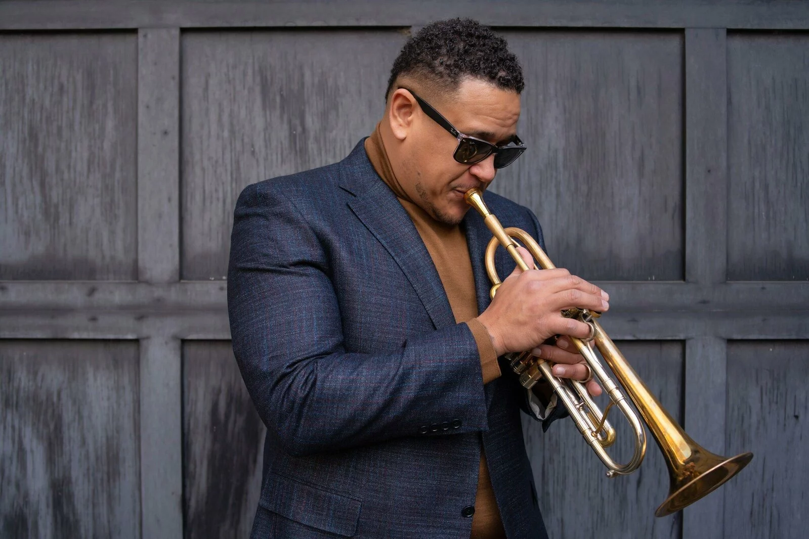 A Black man with dark glasses in a blue blazer blows into a trumpet.