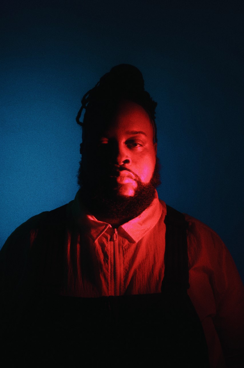 A three-quarter portrait of a well-built man against a blue background who is dramatically lit from his left with a red light