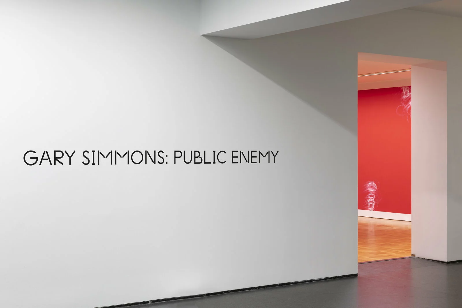 A sliver of a room painted red with white drawings on it is just visible beyond a wall with black text that reads Gary Simmons: Public Enemy.