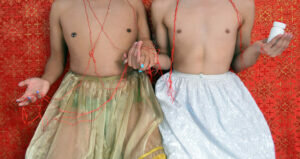 The torsos of two shirtless men holding hands against a red wall. They are wearing slips and are draped in a red thread.