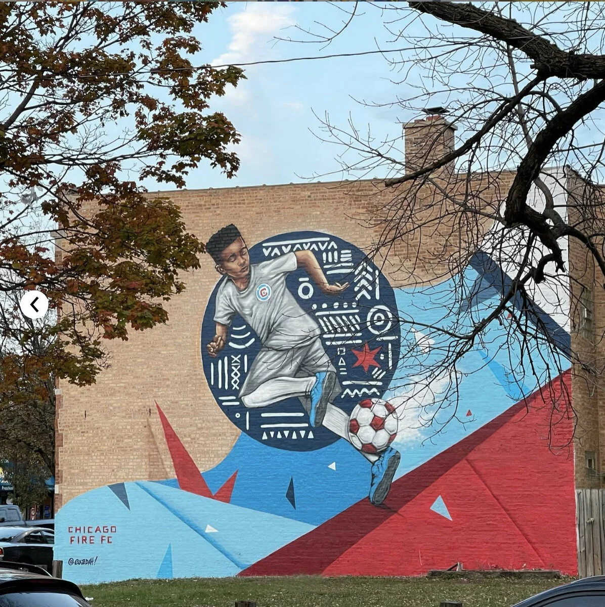 Mural on a brick wall outdoors. It features a young, black male wearing a Chicago Fire FC uniform and kicking a soccer ball. The background consists of a large black circle with linear symbols as well as blue and red bands of color—perhaps a deconstructed version of the Chicago Fire's club logo.