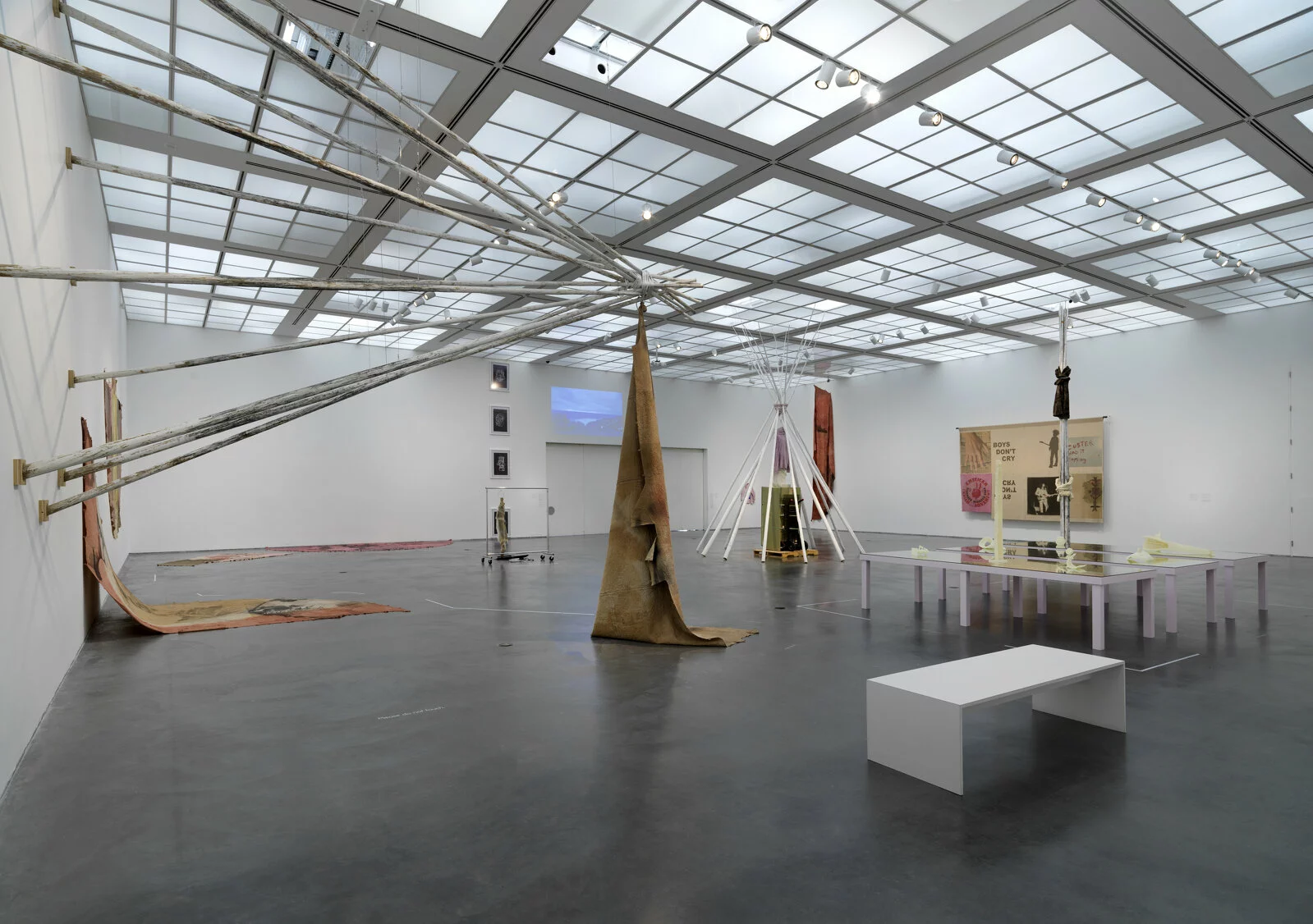 A large gallery space with many artworks installed on the walls and floor, including posts put together in a tipi formation that juts out from the wall.