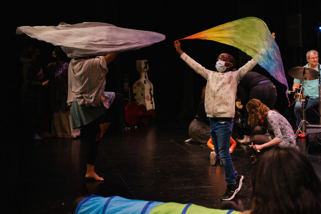 Two people raise large, colorful pieces of fabric over their heads on a darkened stage.