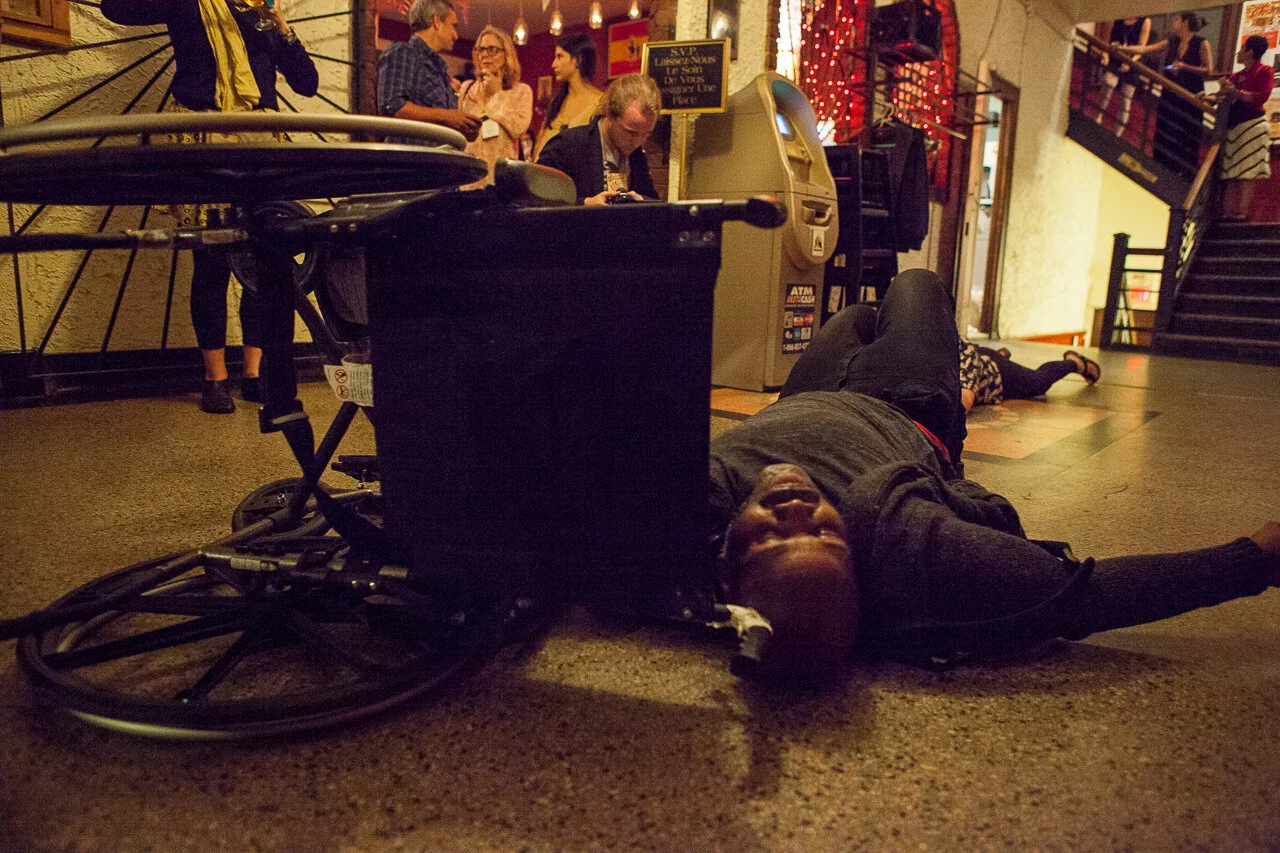 On the landing of a staircase, a brown-skinned large-bodied black person lies on a marble floor next to a manual wheelchair that is turned on its side. In the background is a automated bank machine and a diverse group of people in various positions. Some are looking intently at the person, others are talking with each other.