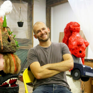 A light-skinned man smiles at the camera in a room amid colorful, biomorphic sculptures.