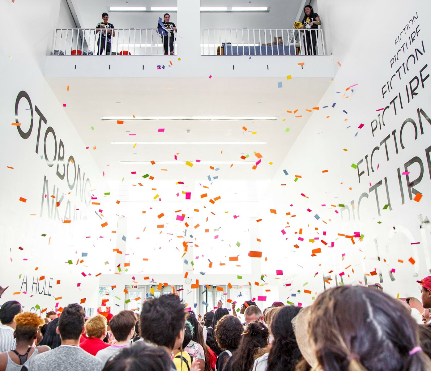 Colorful pieces of paper float over a crowd of people in a large, open white room.