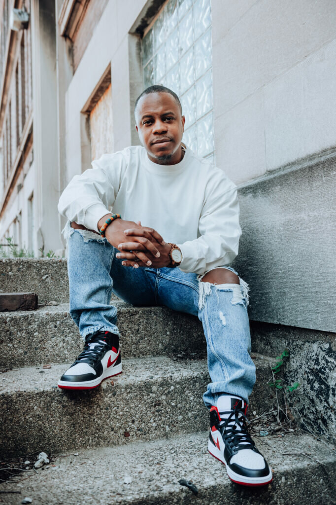A black man wearing a white sweatshirt and jeans poses on a set of concrete steps. His arms rest on his knees, hands loosely clasped together.