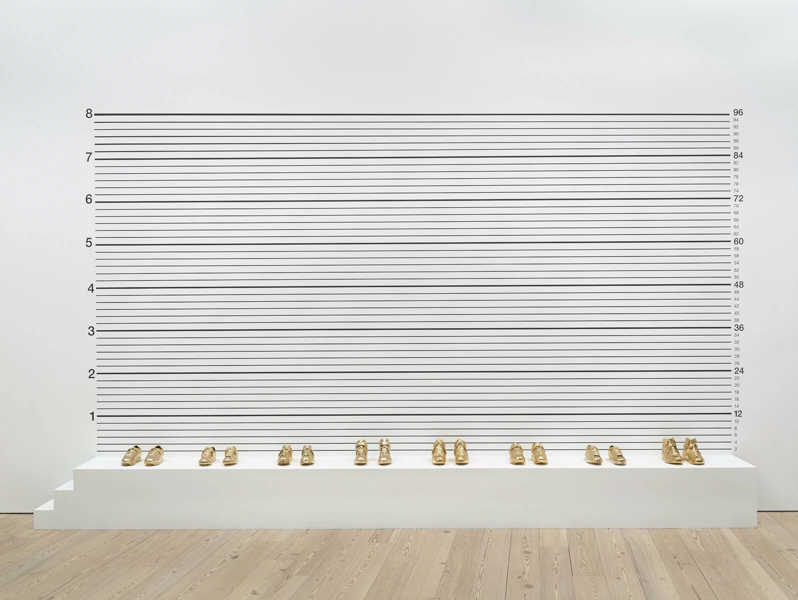 Photo of 8 pairs of gold-plated basketball shoes evenly spaced out on a white platform in front of a white wall with black numbers and lines.