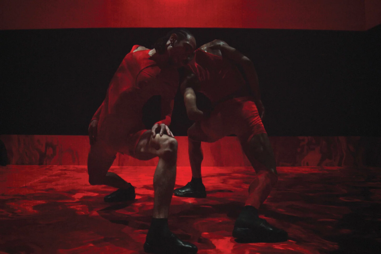 Two Black people wearing red shorts and tops crouch in a wide-legged stance and lean their hands to touch.