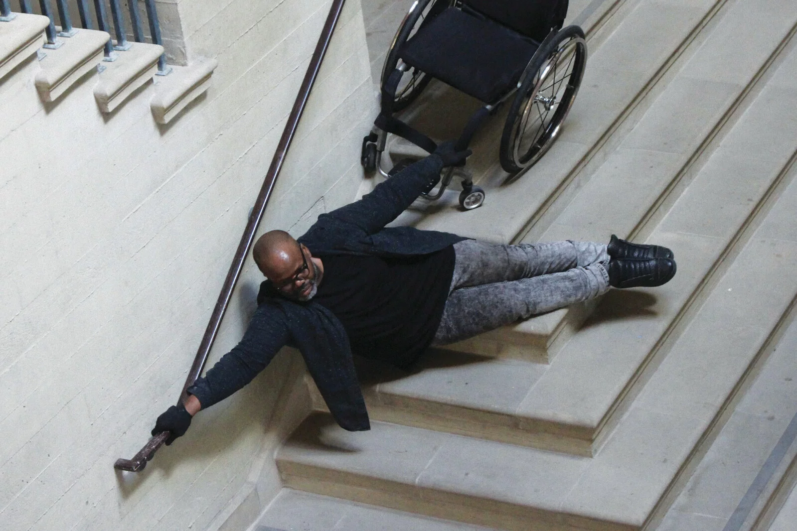 A brown-skinned, large-bodied Black person with a shaved head and beard wearing glasses, a black shirt and grey jeans with one hand on a rail and the other holding a wheelchair, seemingly floating above a set of stairs.