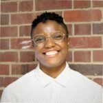 Nic Wyatt is pictured here, standing in front of a brick wall. They are a Jamaican American non-binary individual with short hair smiling at the camera. They are wearing a buttoned polka-dot shirt and gold and silver wire frame glasses.