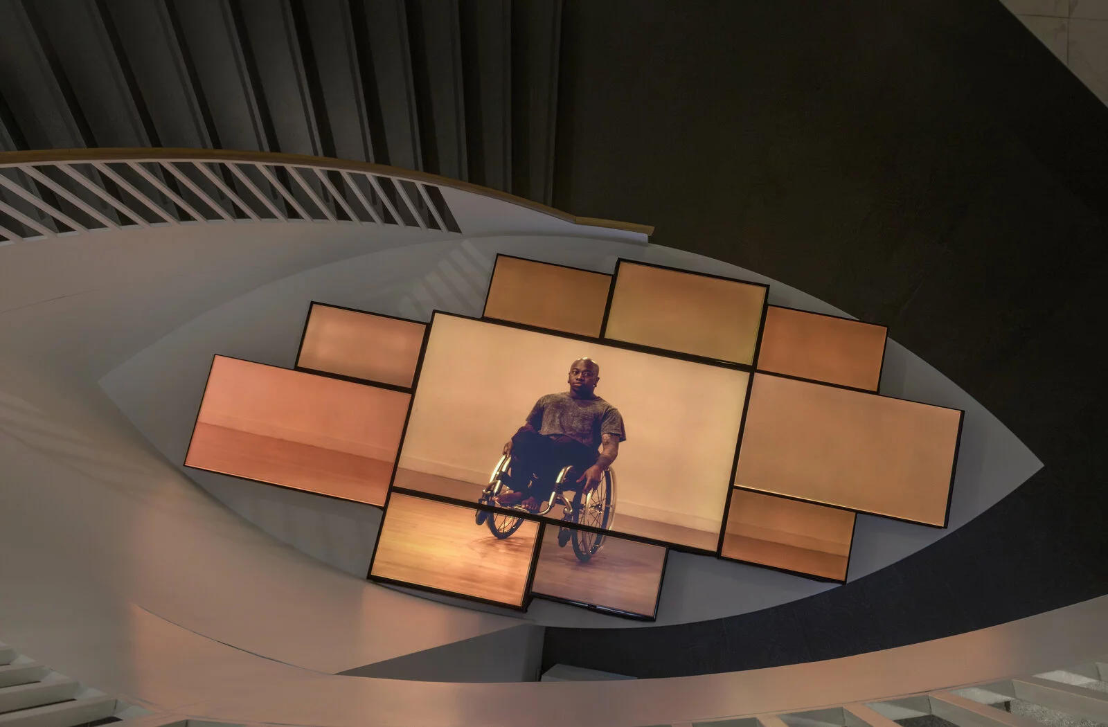 Ten TV screens of varying sizes arranged tightly together display a single image of a person who uses a wheelchair against a peach backdrop.
