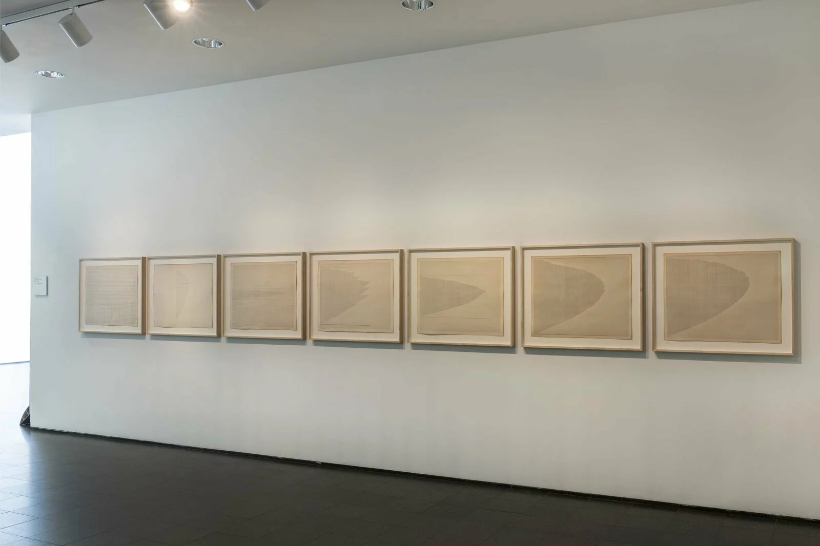 Seven artworks in light wooden frames hang on a white wall in a low-lit space.