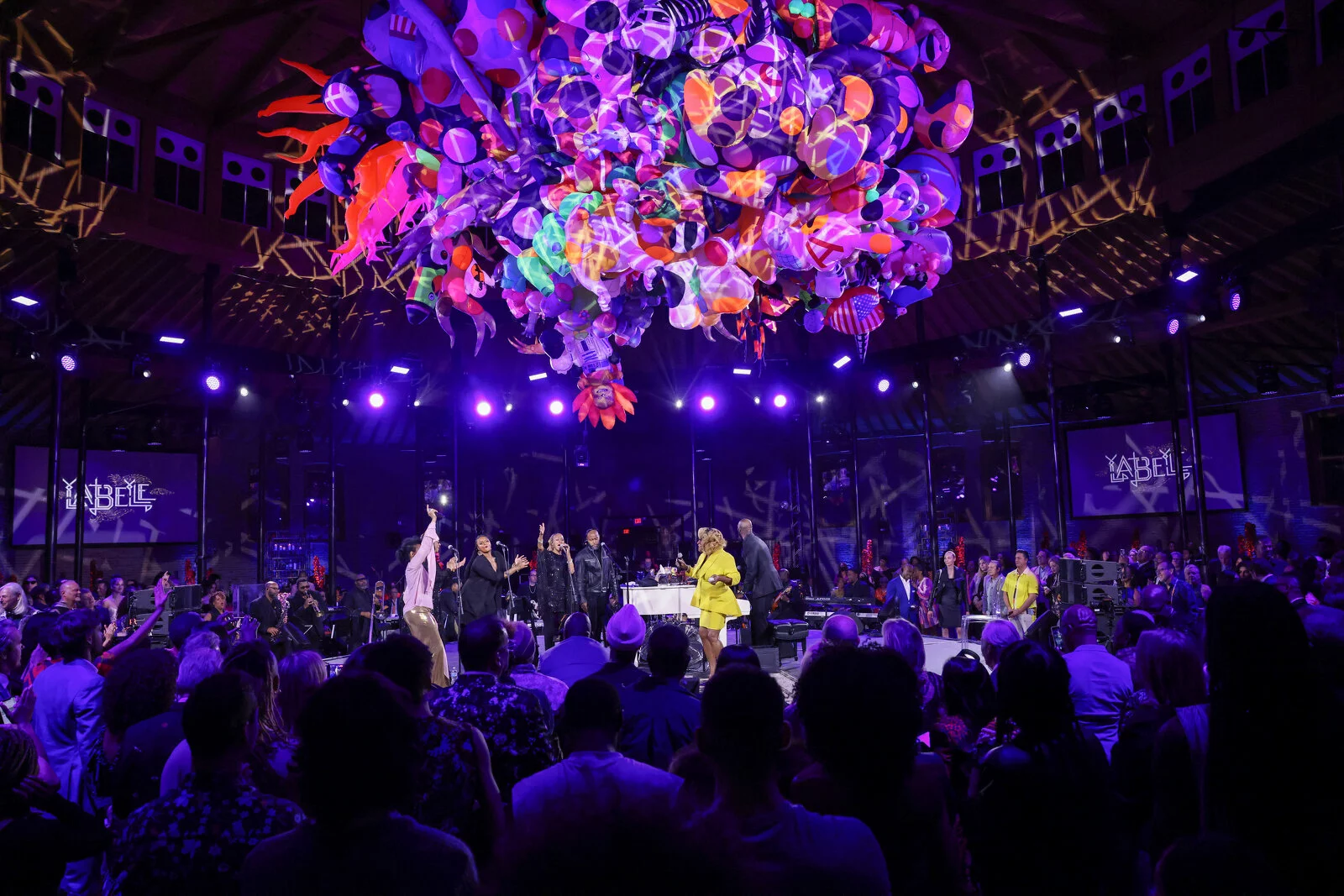 Photo of event with performers onstage with an art installation in purple, red, orange, green, and pink hanging above them.