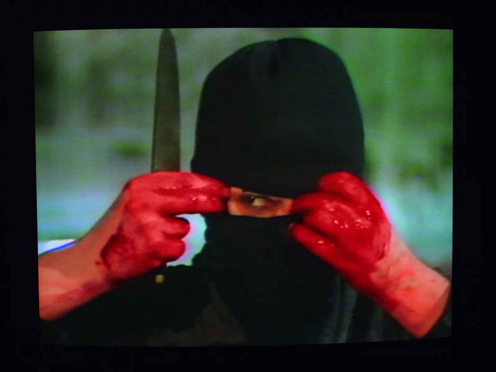 Photograph of person in a black mask holding a knife with bloody hands.