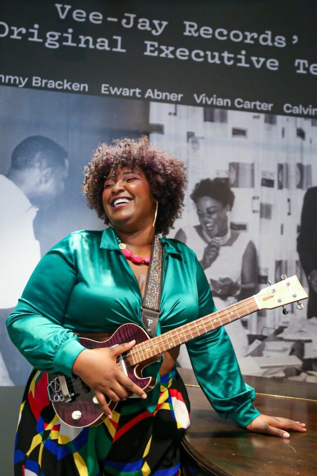 Photo of a brightly dressed Black woman standing in front of black-and-white posters labeled Vee-Jay Records. Her left hand rests on a wooden table while her right hand holds onto a guitar. She smiles broadly towards something off camera.