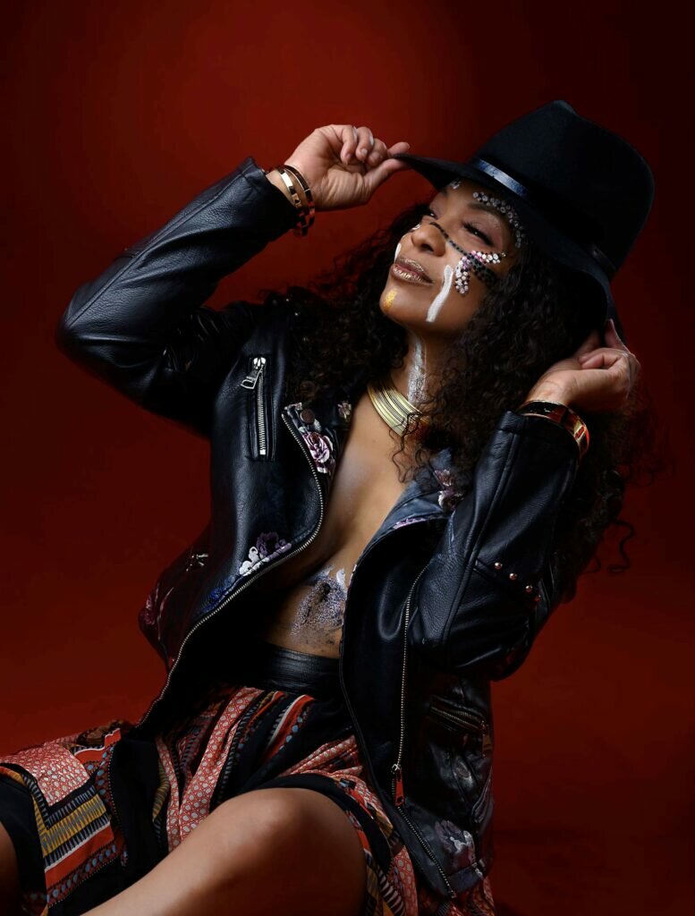 A woman of color poses against a garnet red backdrop. Her face is painted and she wears a black fedora and a leather jacket. Her hands frame her face as she holds onto the front and back of her hat.