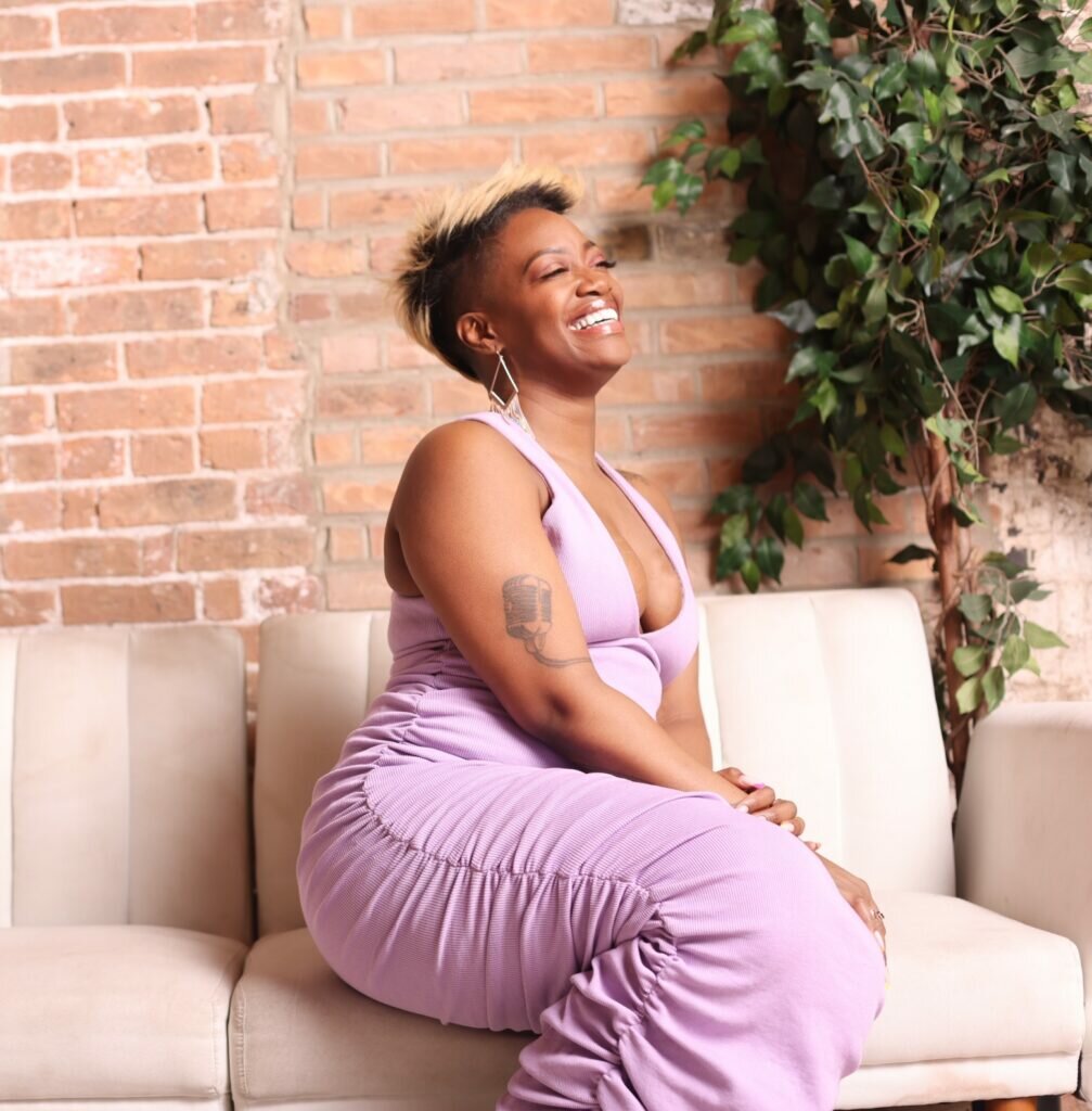 Photo of a Black woman sitting on a couch. She looks up and smiles at something off camera.