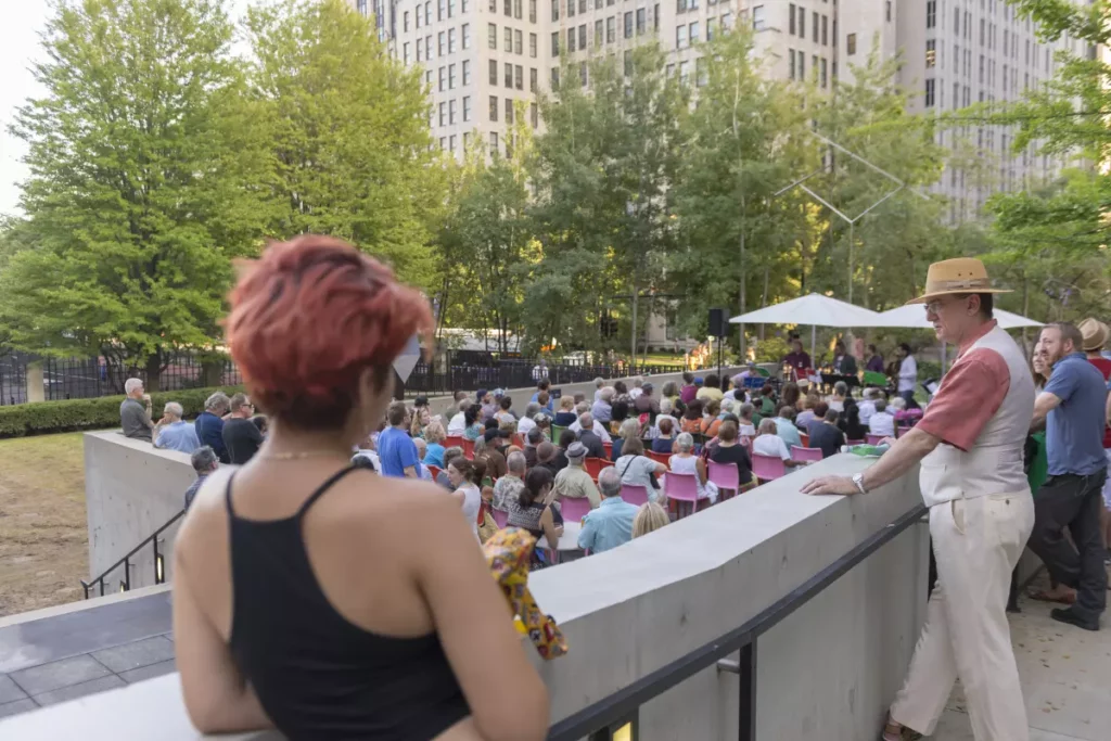 A person with short, pink hair, their back turned away from the camera, looks over a crowd of people. In the background, a band plays.