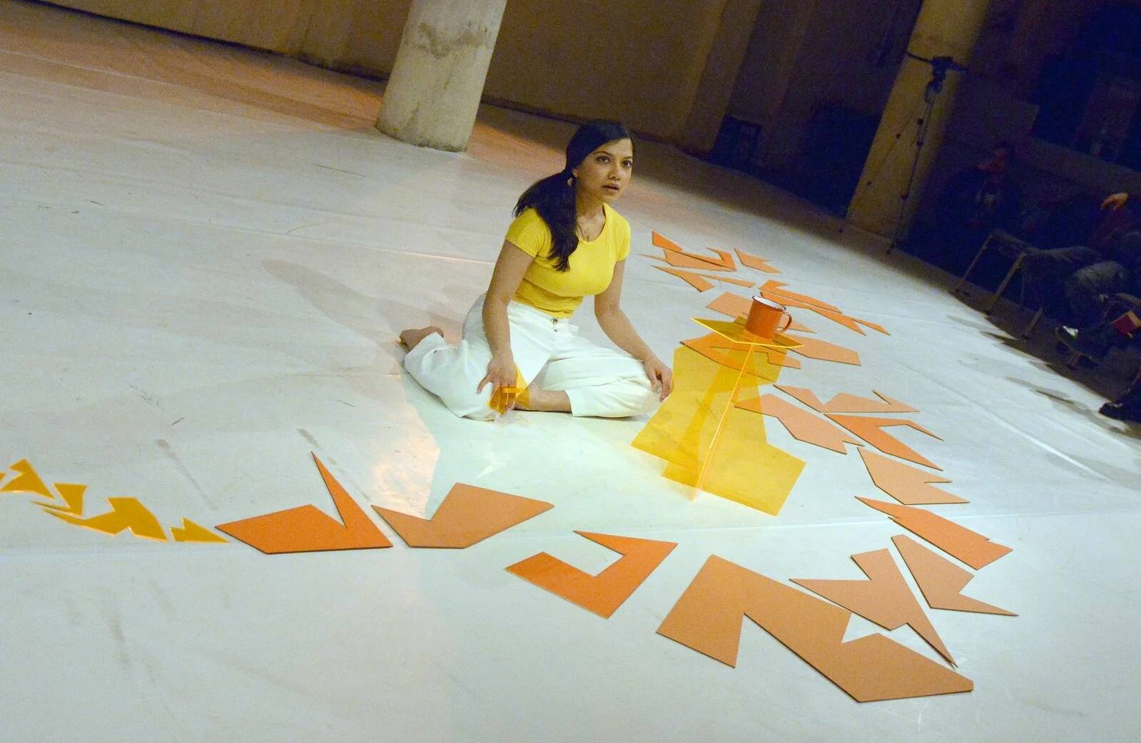 A woman sits on a sleek white floor among large geometric orange paper cutouts.
