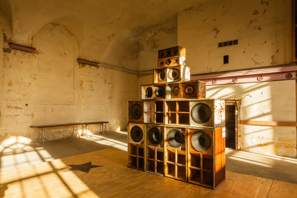 A stackful of various sized speakers in an otherwise empty room. Light streams from the windows, which cast shadows on the speakers, walls, and floor.
