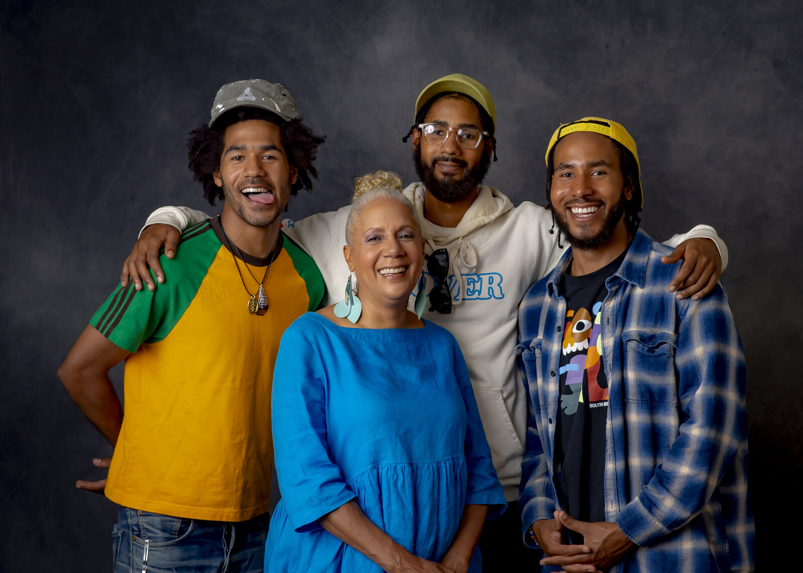 A group portrait of a woman surrounded by three younger men against a gray backdrop.