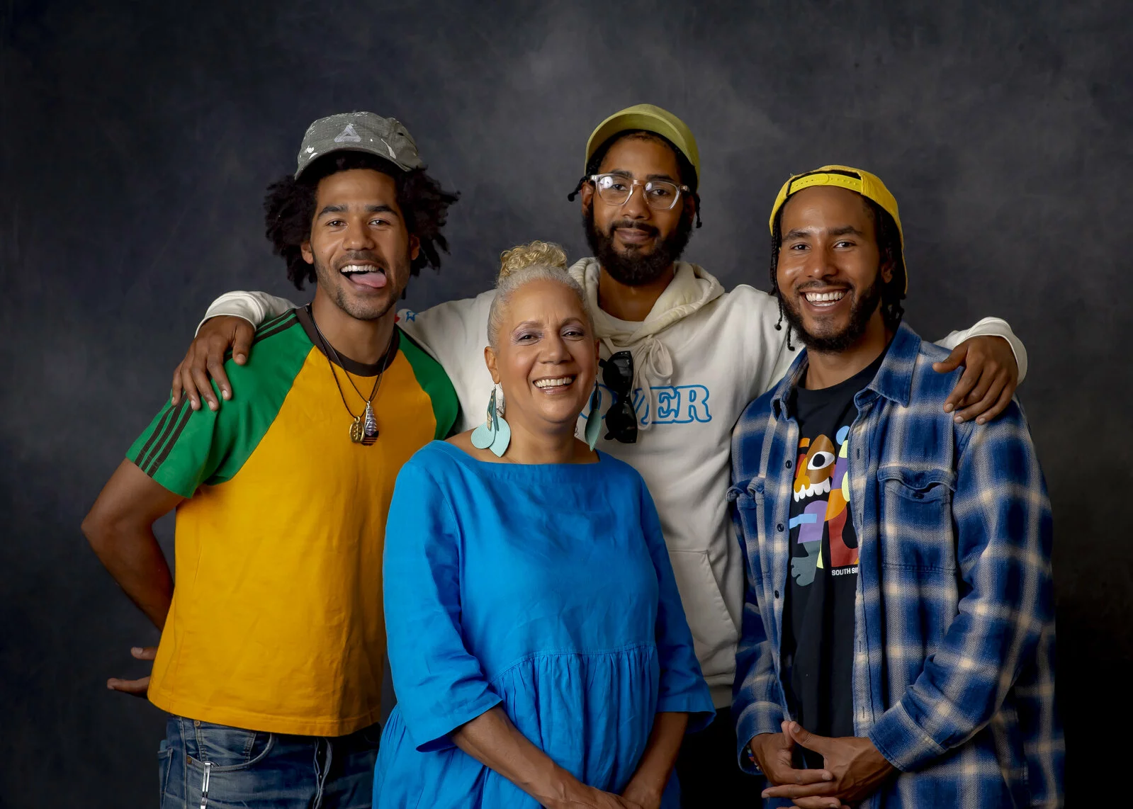 A group portrait of a woman surrounded by three younger men against a gray backdrop.