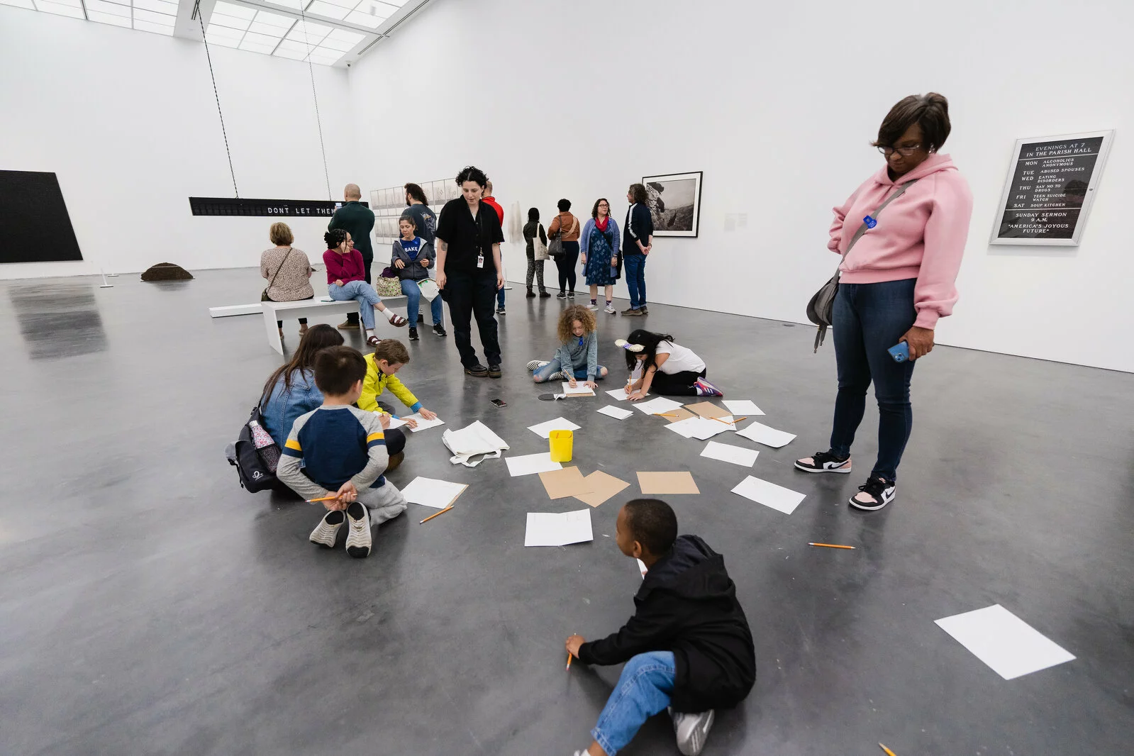A group of kids peruse pages of paper scattered across a gallery floor as adults watch on.