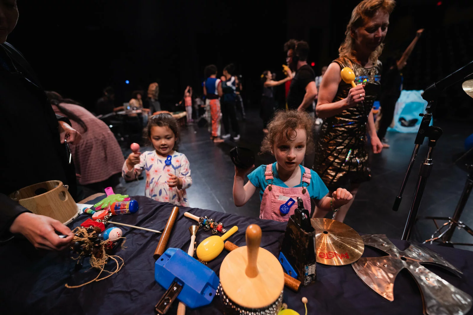 Kids and adults gather around tables filled with various noise making objects on a black stage.