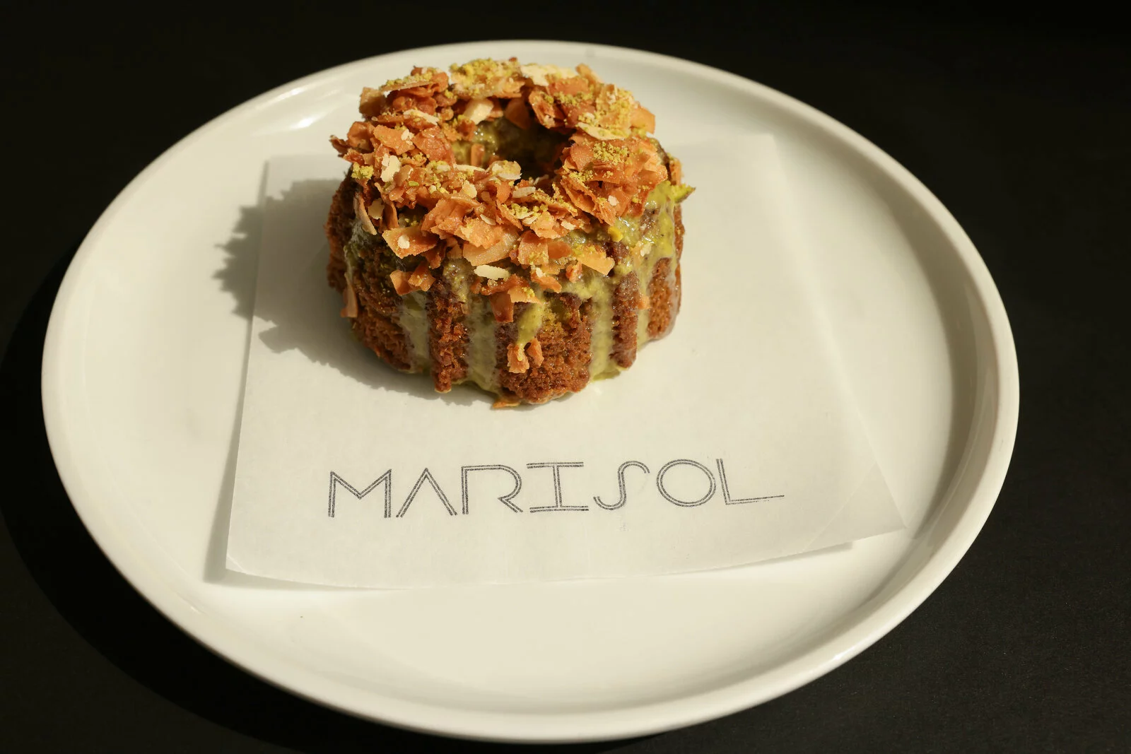 A beautiful, round pastry sits on a parchment paper marked with the word Marisol on top of a white plate against a black background.