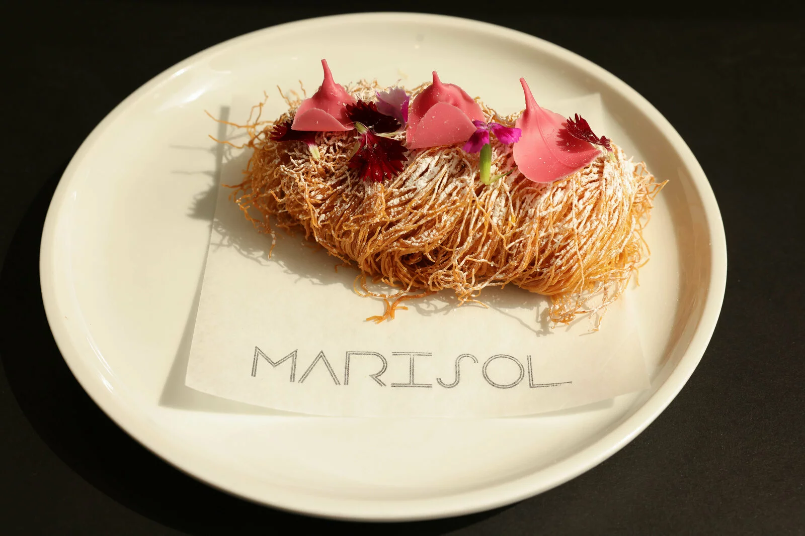 Long strings of shredded dough with flowers on top rests on a parchment paper labeled Marisol on top of a white plate.