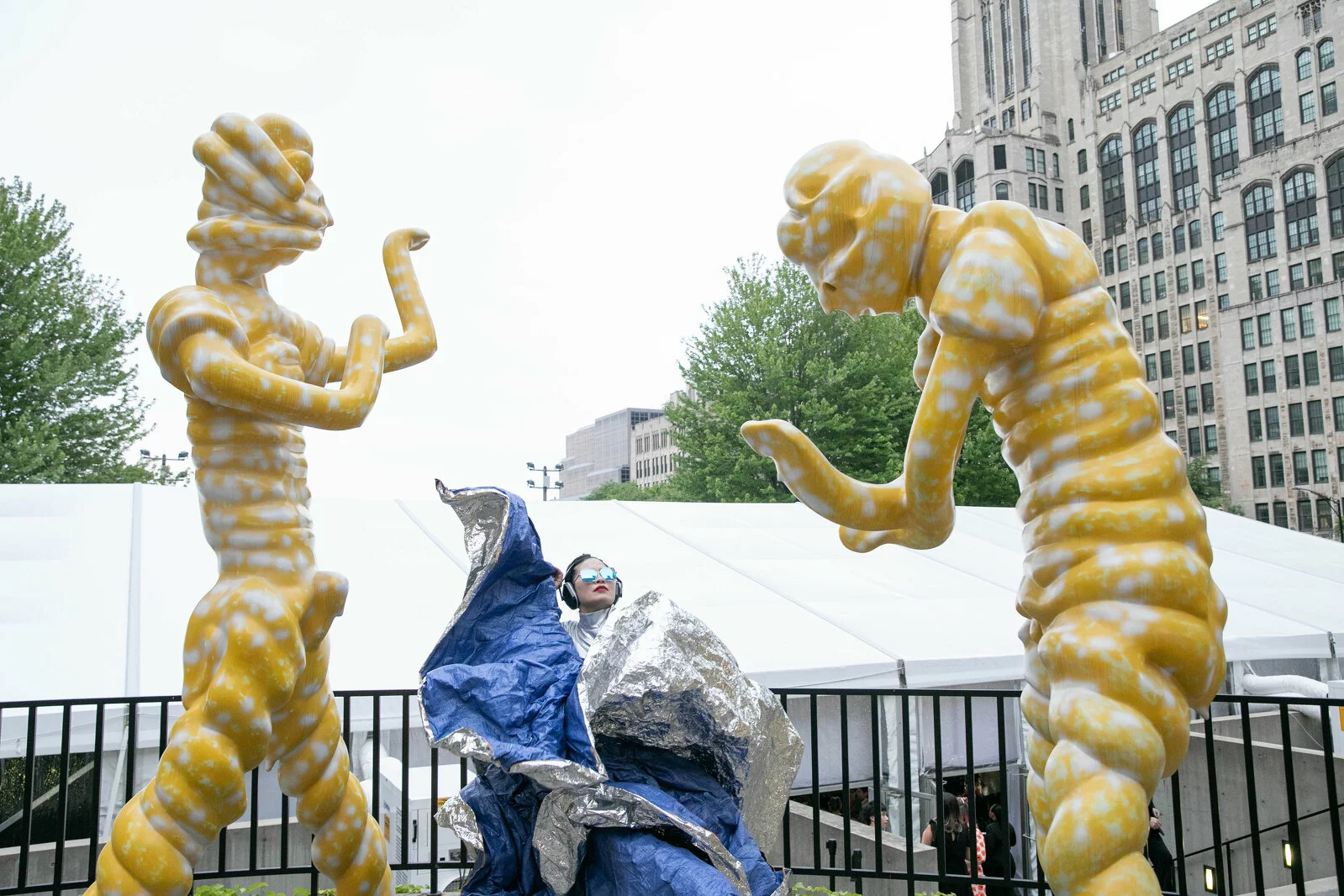 A person in a blue and silver outfit poses flamenco style between two larger-than-life yellow and white sculptures of humanoid figures in different dancing poses.
