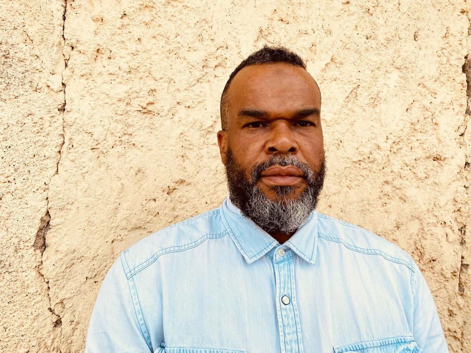 Headshot of Jamal Cyrus, a Black person with a black and white beard, wearing a light blue buttoned up shirt.